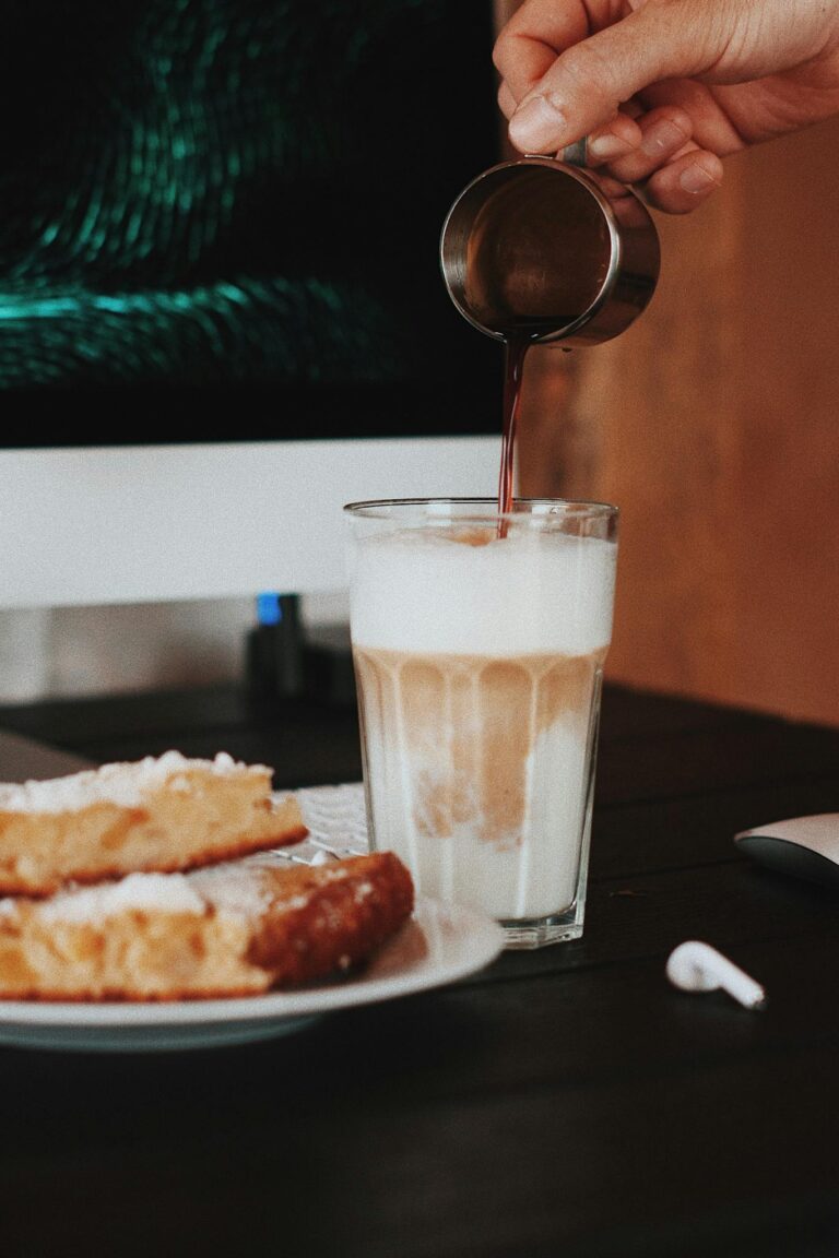 Hand pouring syrup into iced coffee with pastries beside, creating a cozy cafe vibe.